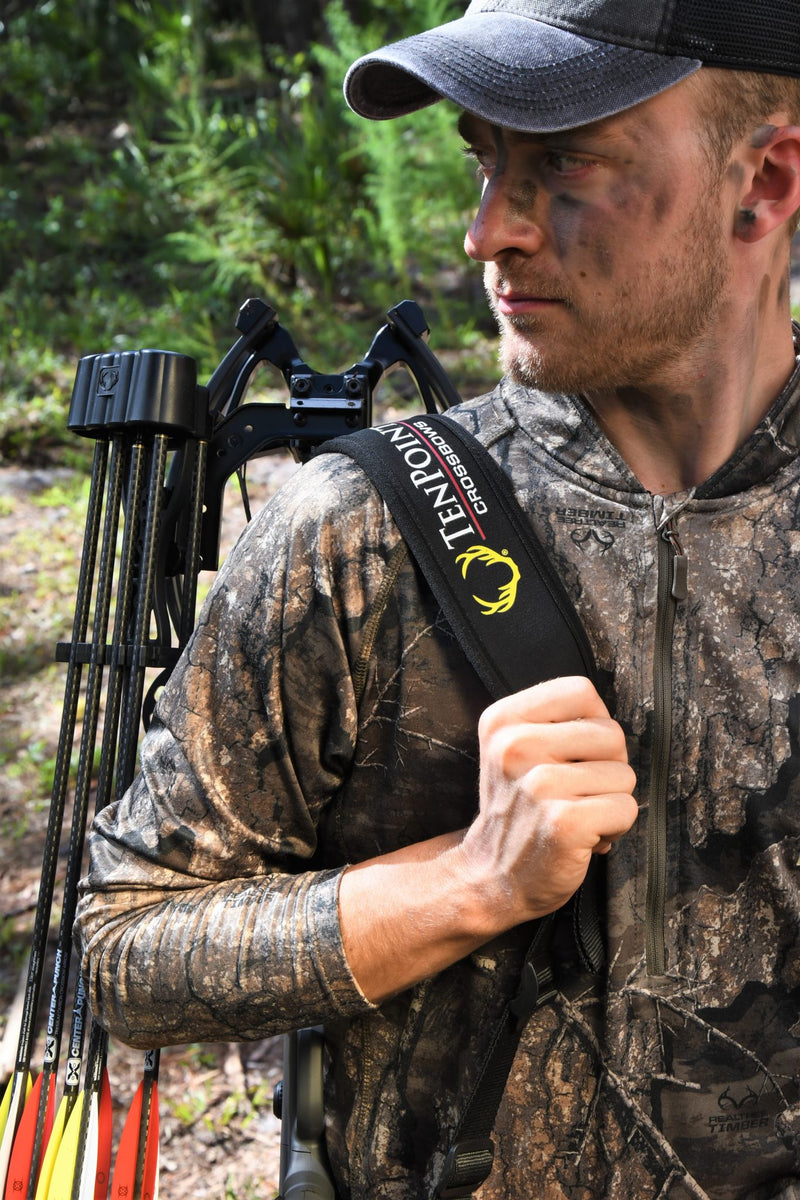 A man in camouflage hunting clothing and a cap carries a TenPoint bow on his back in a forest setting.