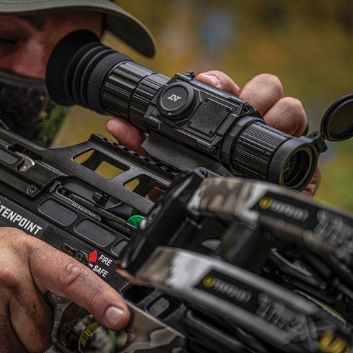 A close-up action shot of a person in camouflage clothing aiming a TenPoint compound bow with an attached black scope, set against a blurred outdoor background.